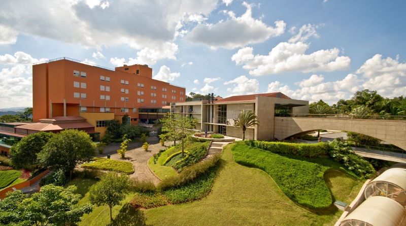 Vista panorâmica do Almenat Embu das Artes, Tapestry Collection by Hilton, com fachada de tons terrosos, jardins tropicais, amplo gramado verde e céu azul com nuvens, hotel de lazer e corporativo a 35 km de São Paulo.