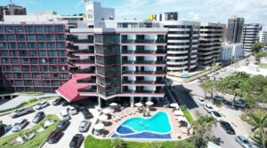 Vista aérea da fachada do Maceió Mar Hotel, edifício branco com varandas e detalhes vermelhos, piscina azul em formato curvo com espreguiçadeiras e guarda-sóis na área externa, estacionamento movimentado e prédios ao redor.