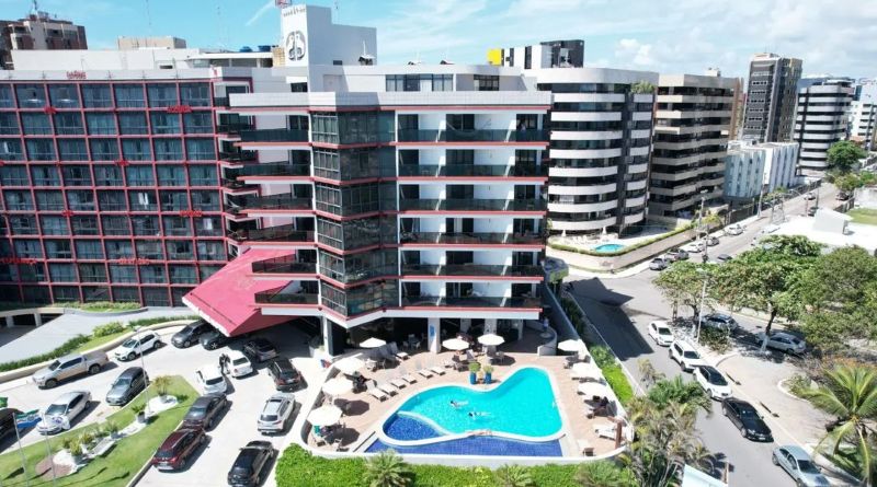 Vista aérea da fachada do Maceió Mar Hotel, edifício branco com varandas e detalhes vermelhos, piscina azul em formato curvo com espreguiçadeiras e guarda-sóis na área externa, estacionamento movimentado e prédios ao redor.