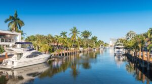 Vista de canal em Fort Lauderdale, Flórida, com iates e barcos ancorados em docas de madeira, palmeiras tropicais nas margens, casas à beira d'água e edifícios ao fundo sob céu azul, destino de luxo e passeios de barco na Flórida.