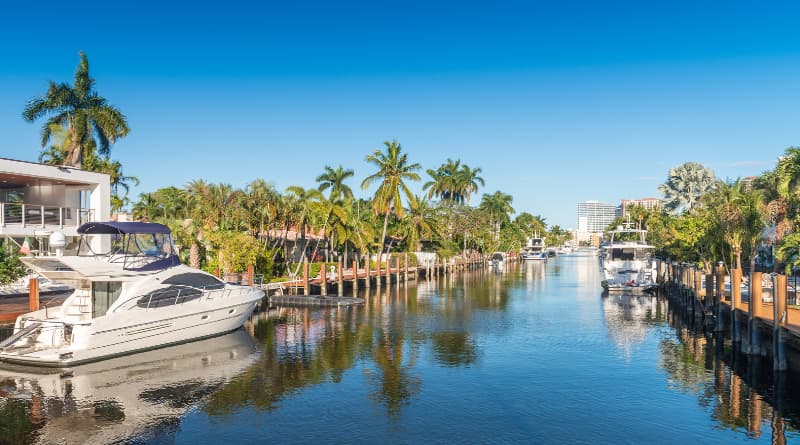 Vista de canal em Fort Lauderdale, Flórida, com iates e barcos ancorados em docas de madeira, palmeiras tropicais nas margens, casas à beira d'água e edifícios ao fundo sob céu azul, destino de luxo e passeios de barco na Flórida.