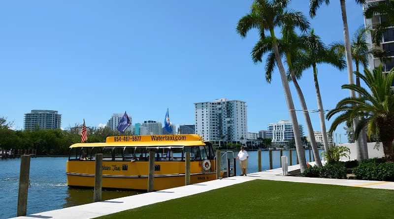 Water Taxi amarelo de Fort Lauderdale navegando por canal com passageiros a bordo, atracado em píer com palmeiras e gramado à beira d'água, arranha-céus e skyline da cidade ao fundo sob céu azul, transporte turístico aquático na Flórida.