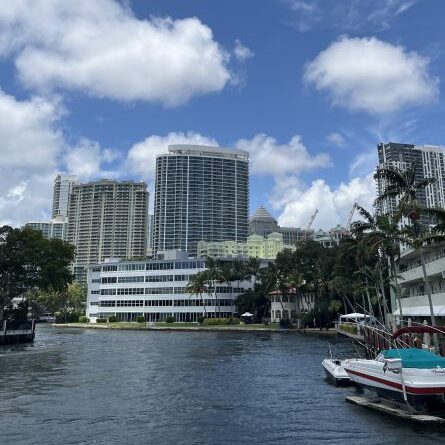 Passeio de Water Taxi pelo canal de Fort Lauderdale com vista para o skyline da cidade, arranha-céus modernos e edifícios à beira d'água refletidos no canal, palmeiras nas margens, barcos atracados e céu azul com nuvens, transporte turístico aquático na Flórida.